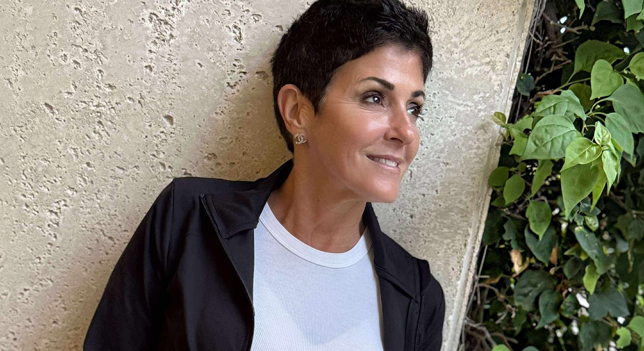 Woman smiling against a textured wall with greenery.